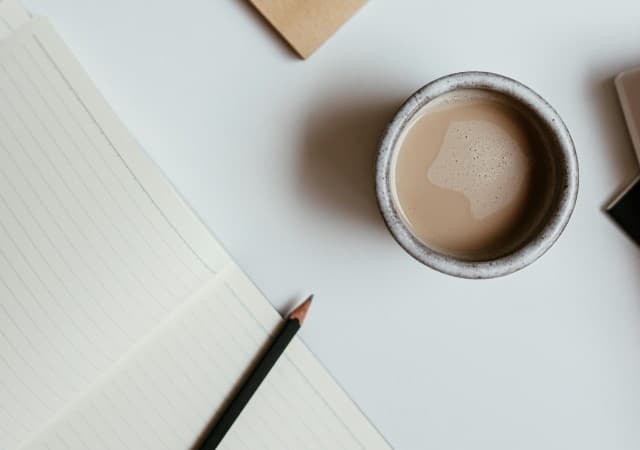 A lined notebook with a pencil on top of it. Also, contains a cup of coffee on the side.